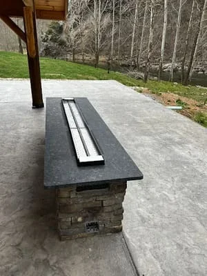 Stone veneer outdoor kitchen island with dark countertop and built-in grill on concrete patio