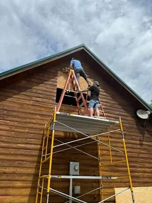 Worker installing wood siding on gable end with yellow scaffolding and safety equipment